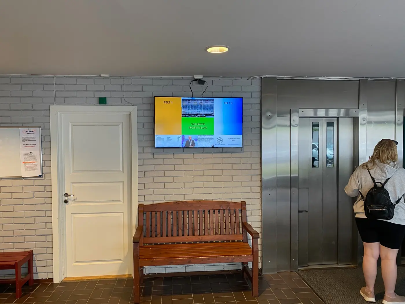 Infoskjerm på teglvegg ved heisen, med benk og dør